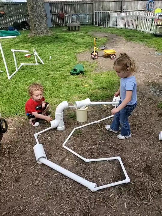 Two children working together to build a run of white pipes in the backyard play area