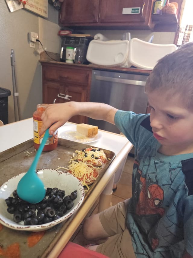 A child spooning black olives onto homemade mini pizzas
