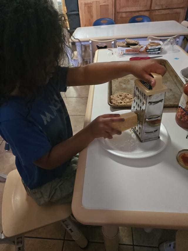 A child grating cheese onto a plate while making mini pizzas