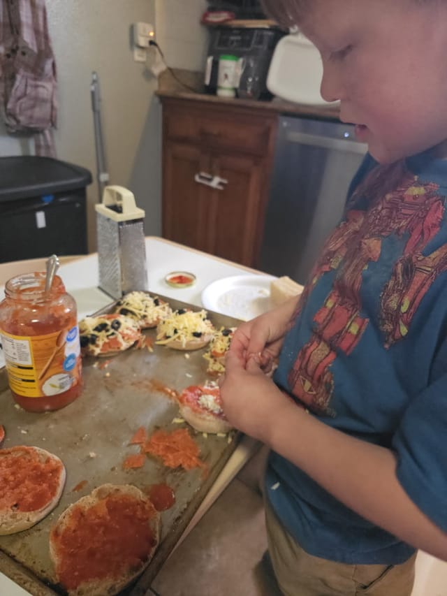 A child spreading sauce and cheese onto English-muffin pizzas